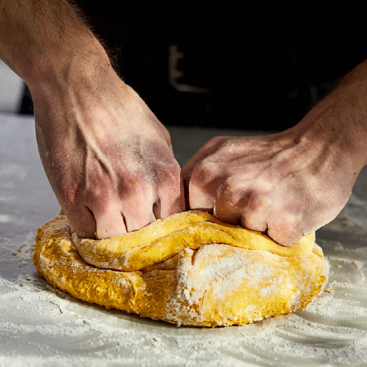 Mani che impastano un impasto di colore giallo su una superficie infarinata, preparando pane o pasta per dolci.
