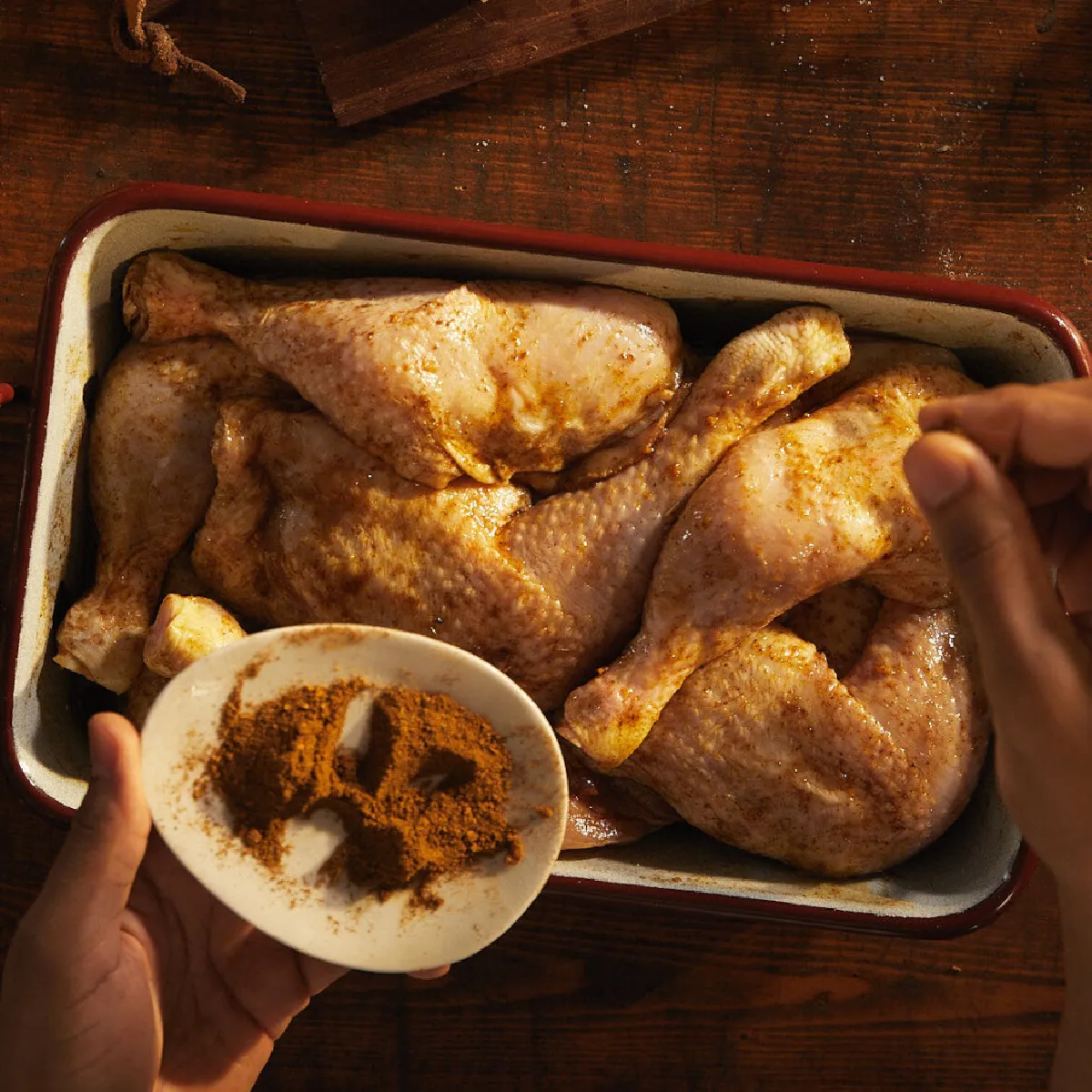 Mani che cospargono una miscela di spezie sui cosci di pollo crudi in una teglia, pronti per essere insaporiti e cotti.
