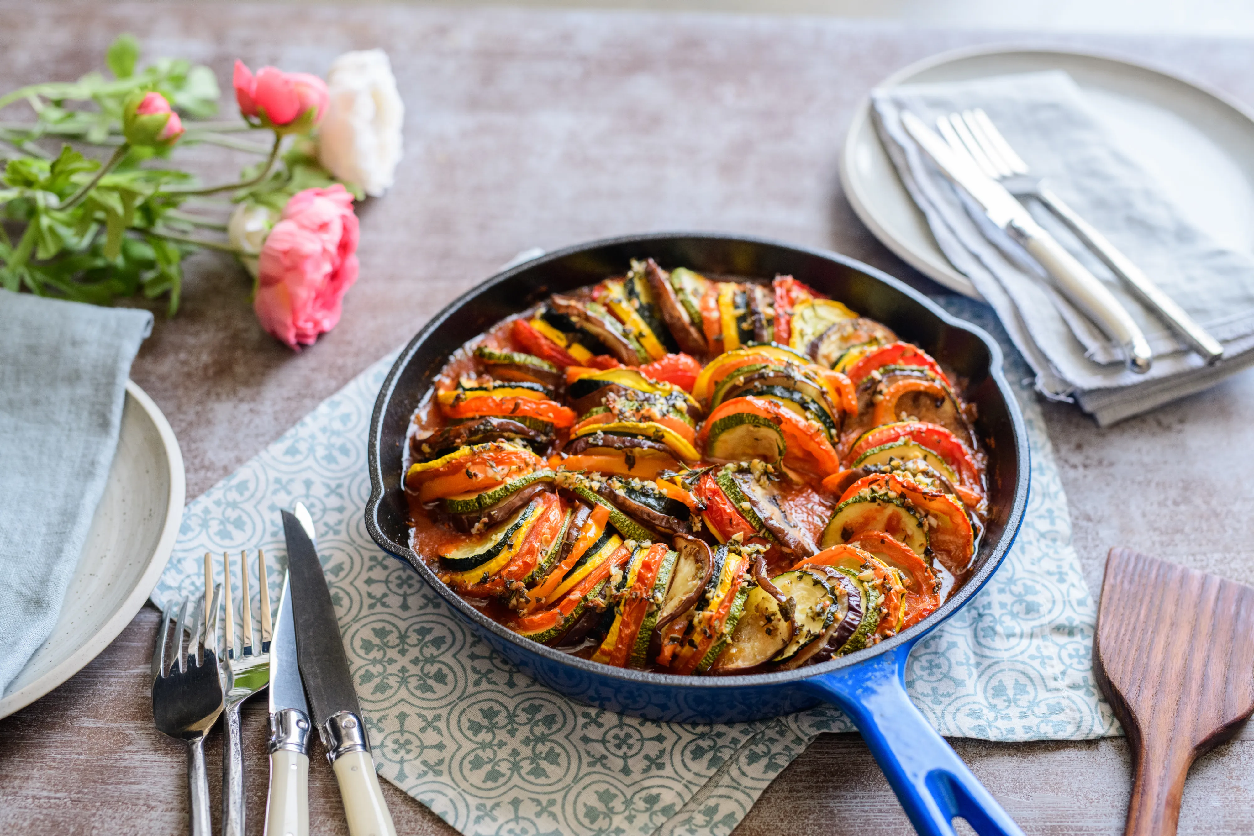 Colorful ratatouille with layered vegetables in a blue skillet, set on a table with plates, cutlery, and pink flowers nearby.
