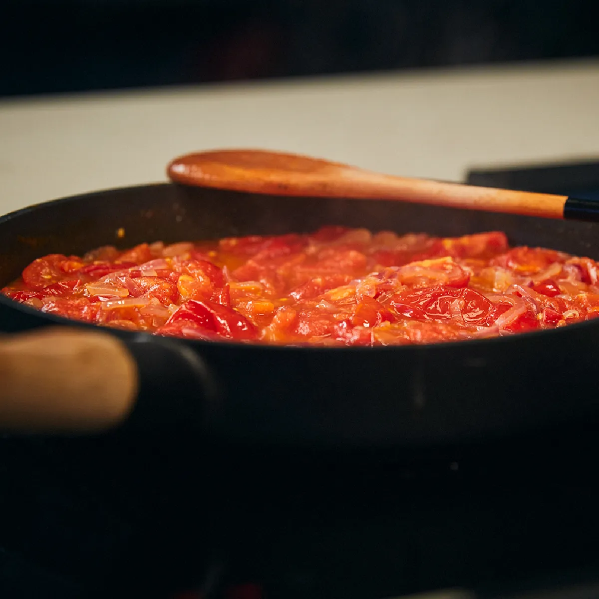 Salsa di pomodoro bollente in una padella di ghisa su un piano cottura, con un cucchiaio di legno che mescola la salsa.