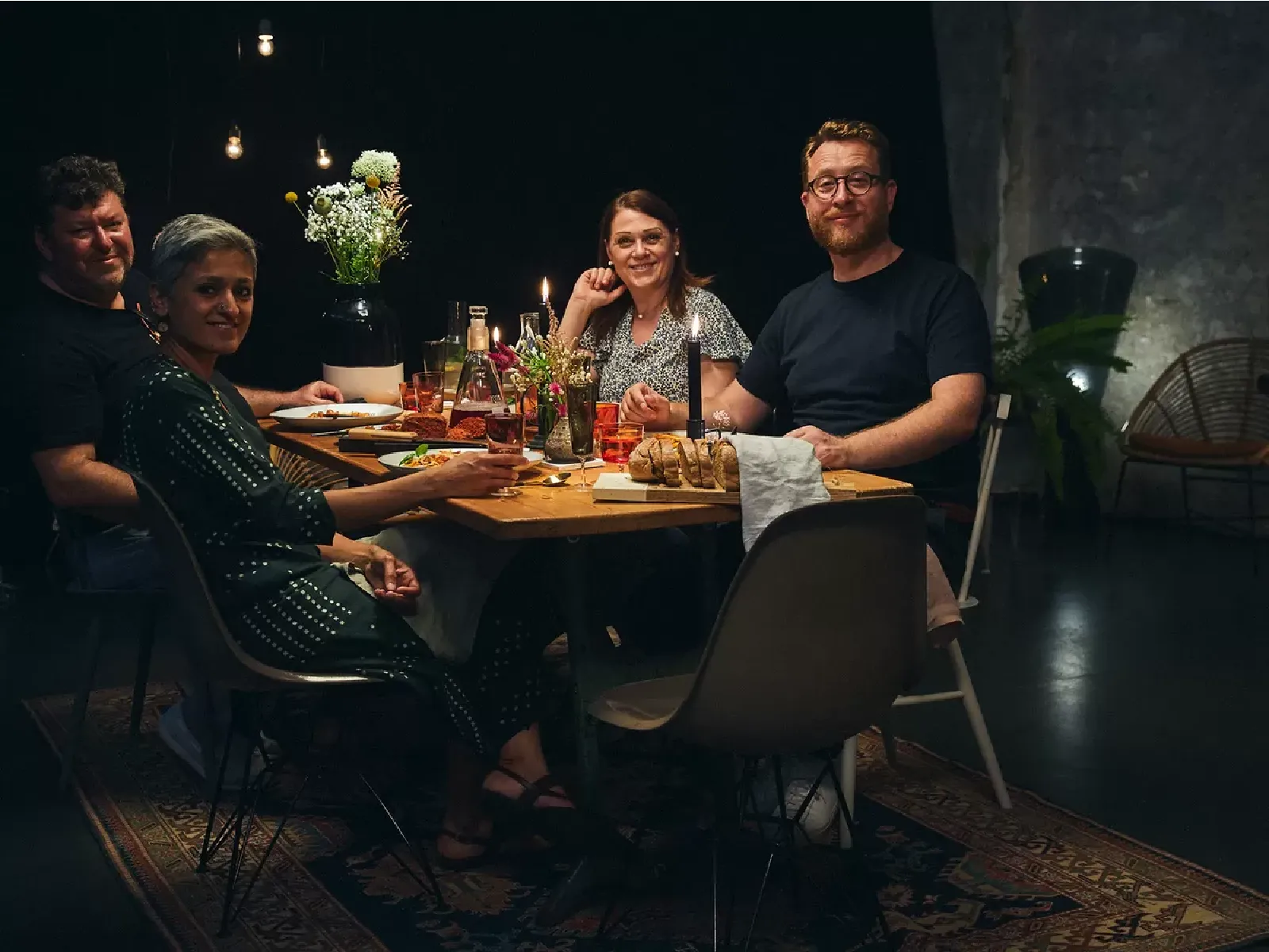 Four people sit around a dimly lit table enjoying a dinner of bread, drinks and floral centerpieces in a cozy and intimate setting.
