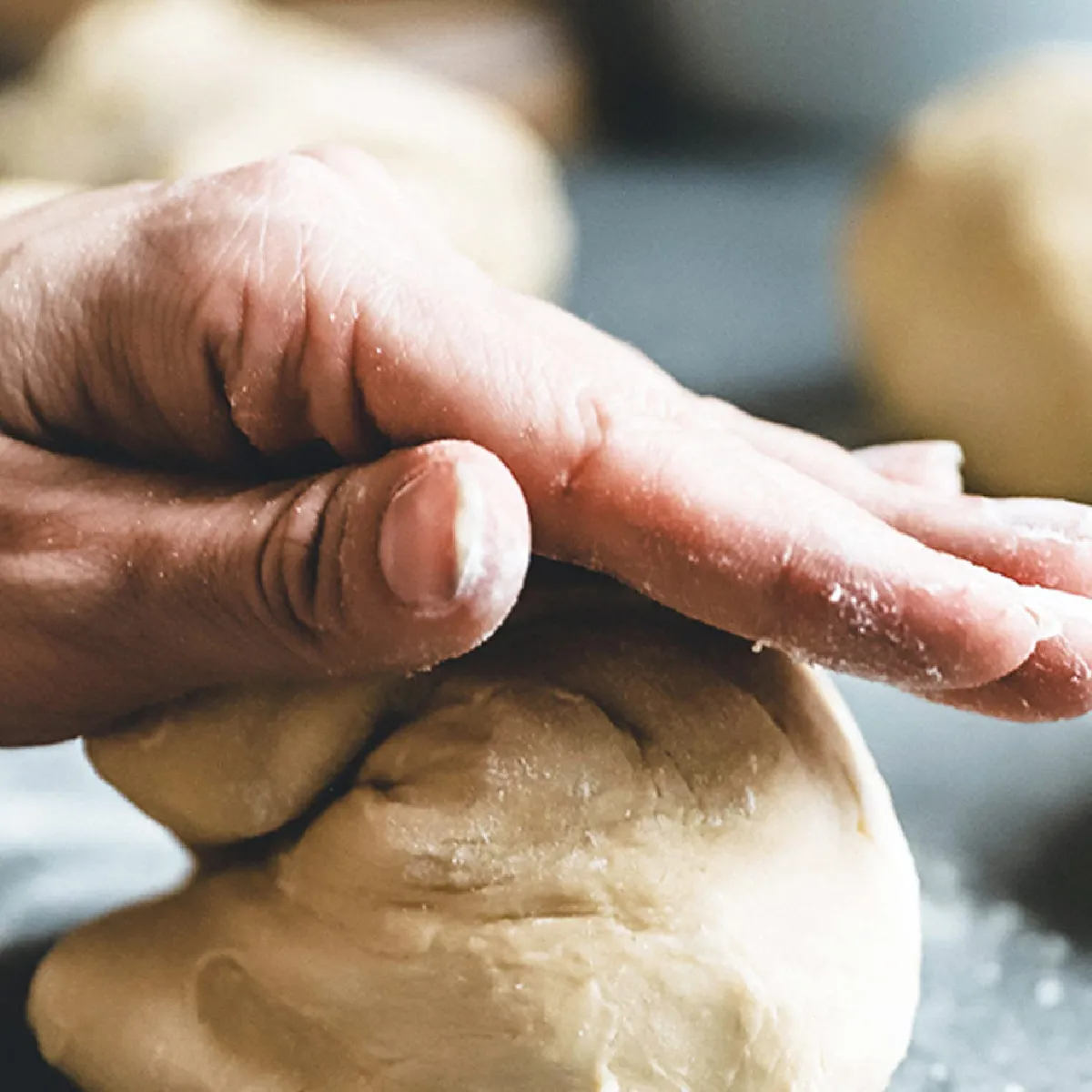 Due mani che reggono una ciotola bianca contenente impasto lievitato, con un cartone di uova e un piano di lavoro sfocato sullo sfondo.