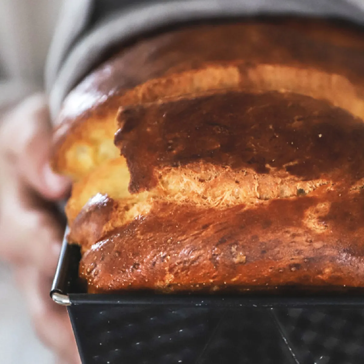 Primo piano di due mani che mostrano uno stampo scuro per pane contenente un filone dorato, lucido e ben lievitato con la crosta fessurata.