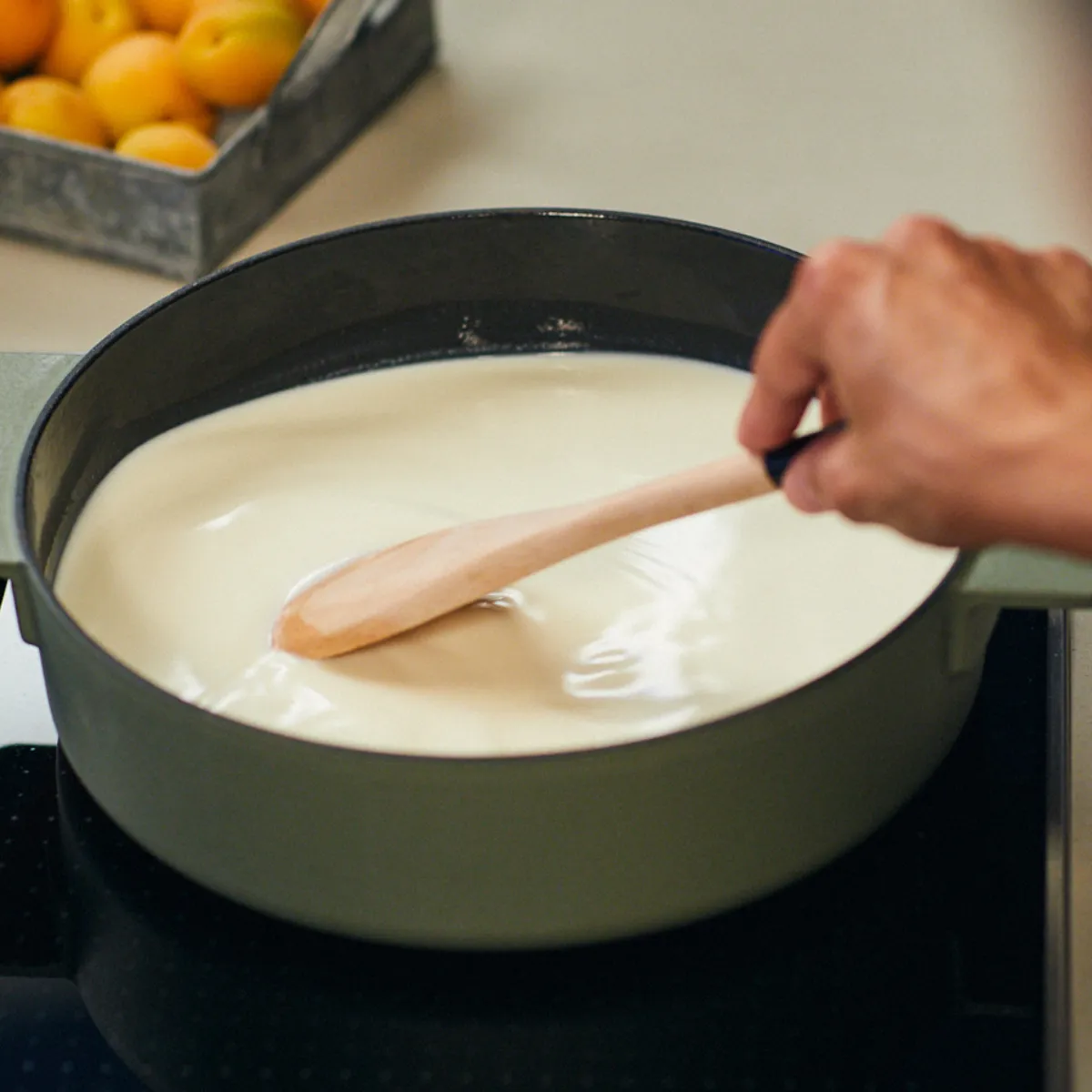 Mani mescolando una pentola di liquido cremoso su un piano cottura, con una scatola di arance visibili sullo sfondo.
