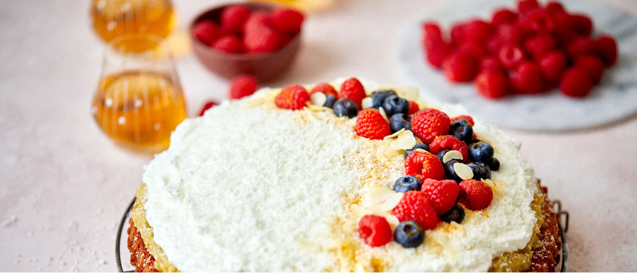 Round cake topped with white frosting, fresh raspberries, blueberries, almond slices, and powdered sugar on a cooling rack.