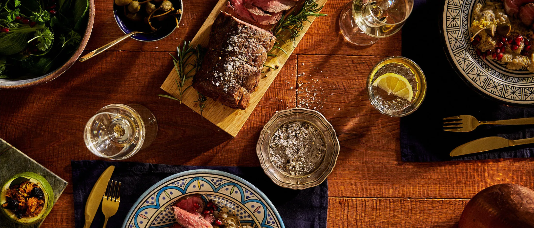 Tavolo rustico con vari piatti, utensili e bevande, tra cui un piatto di carne, insalata e bicchieri di vino.