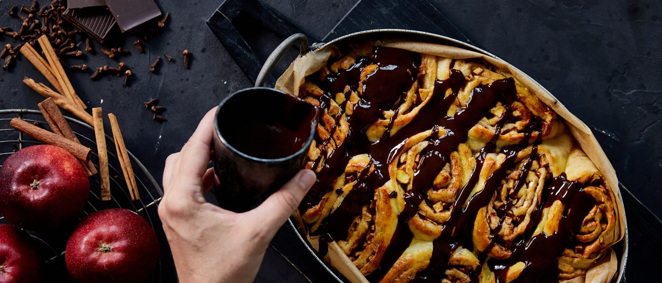 Versare a mano la salsa al cioccolato sui cinnamon rolls appena sfornati in una teglia, con mele, bastoncini di cannella, chiodi di garofano e pezzetti di cioccolato accanto.
