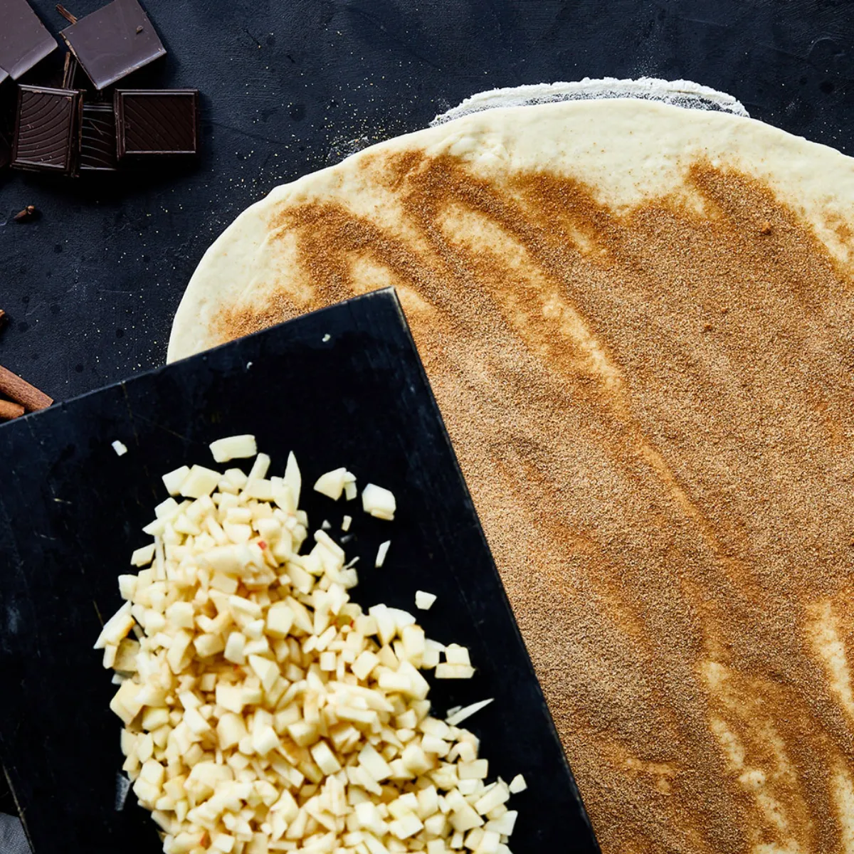 Una sfoglia stesa cosparsa di cannella, accanto a cubetti di mela che vengono versati da un tagliere, con cioccolato, bastoncini di cannella, mele e uova lì vicino su una superficie scura.