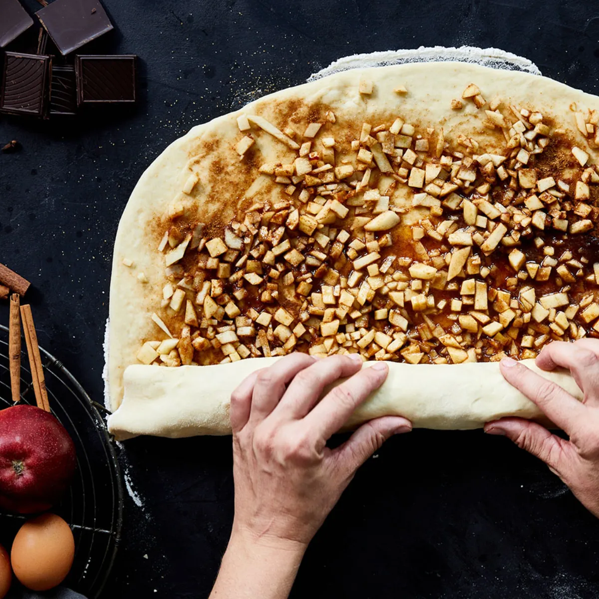 Vista dall'alto di mani intente a stendere una sfoglia rettangolare farcita con mele tagliate a pezzetti, zucchero alla cannella e sciroppo, mentre preparano un dolce alle mele fatto in casa o uno strudel su una superficie rustica scura, circondate da mele rosse fresche, mele affettate, bastoncini di cannella, uova, pezzetti di cioccolato e ingredienti da forno.