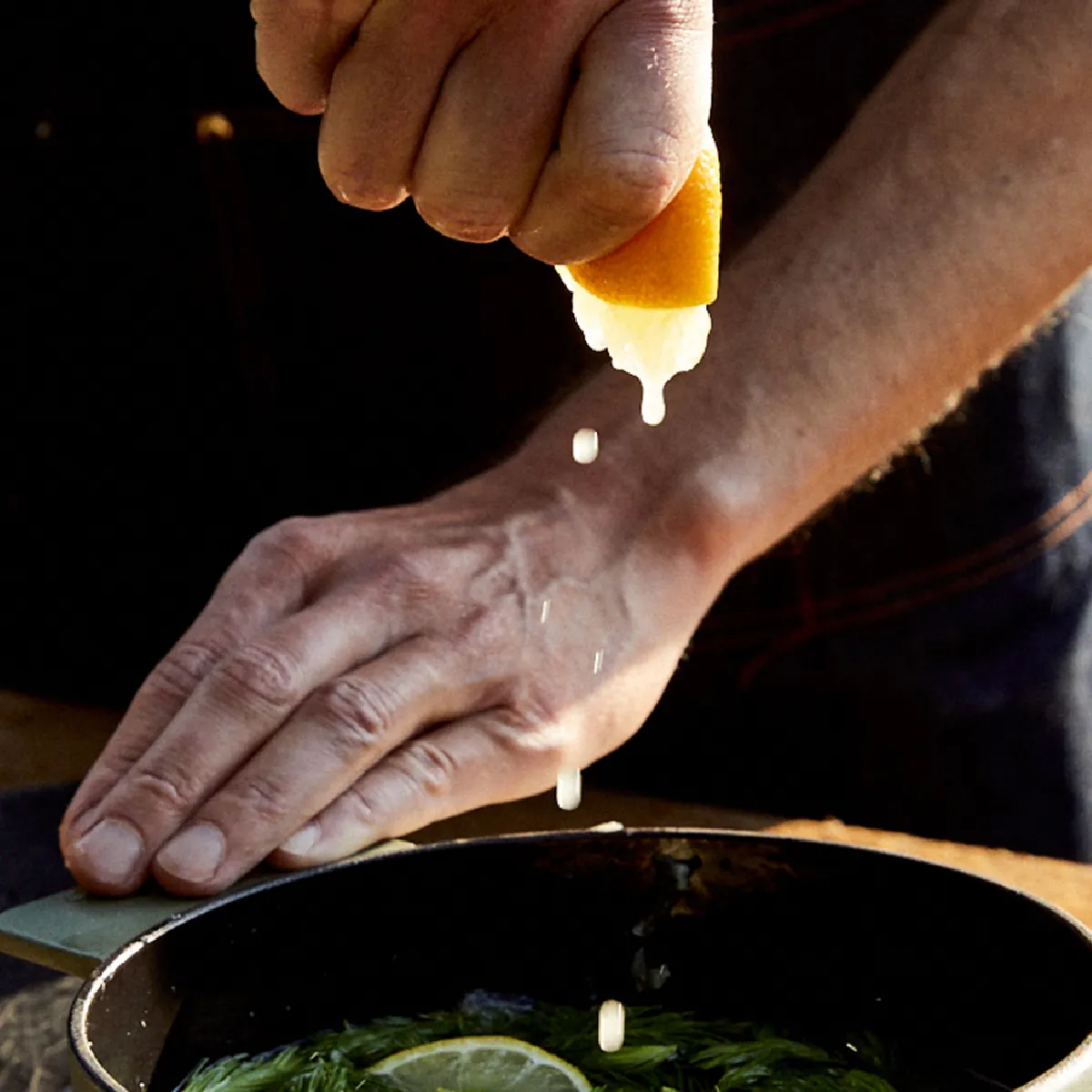 Primo piano di mani che spremono mezzo limone su una padellina con punte di abete verdi e una fetta di lime su un tavolo di legno.