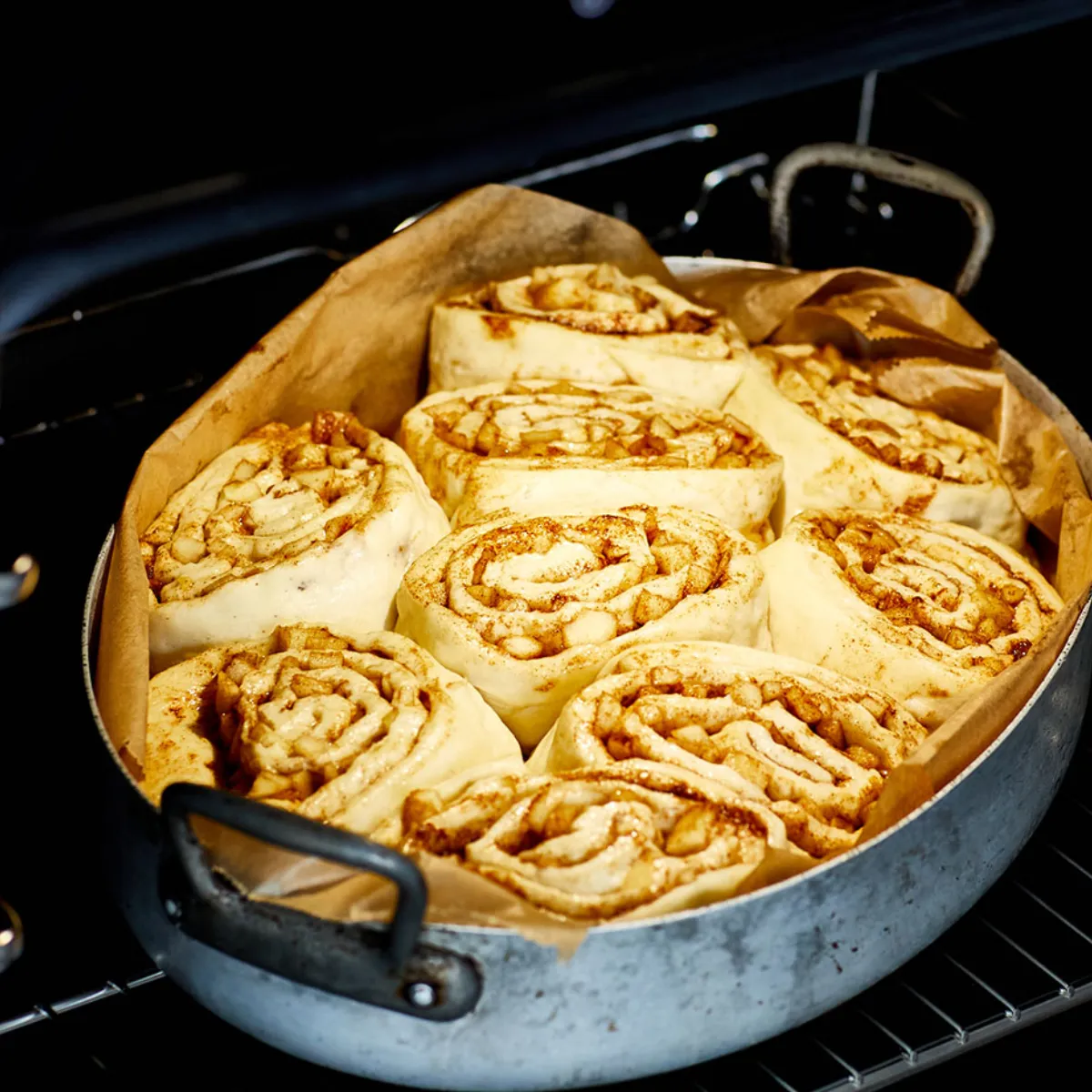 Primo piano di rotolini alla mela e cannella crudi, disposti in una teglia rotonda di metallo rivestita di carta da forno, collocata su una griglia all'interno di un forno; si vede l'impasto a spirale ripieno di zucchero alla cannella e mele a cubetti, pronto per essere infornato in una cucina domestica.