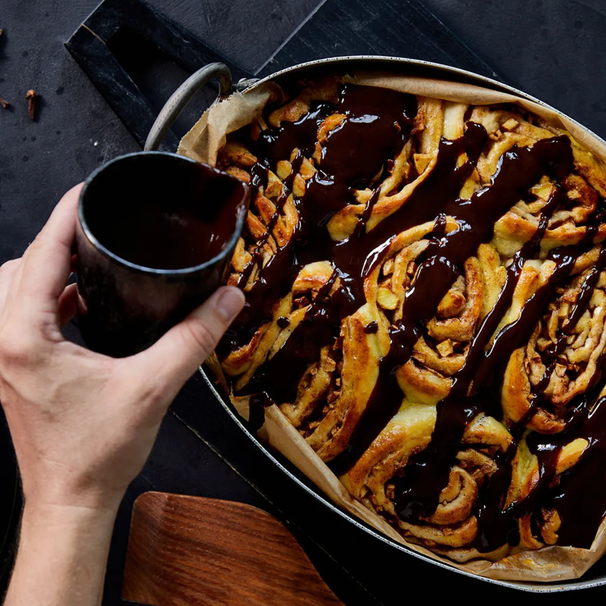 Versare a mano la salsa al cioccolato sui cinnamon rolls appena sfornati in una teglia, con mele, bastoncini di cannella, chiodi di garofano e pezzetti di cioccolato accanto.