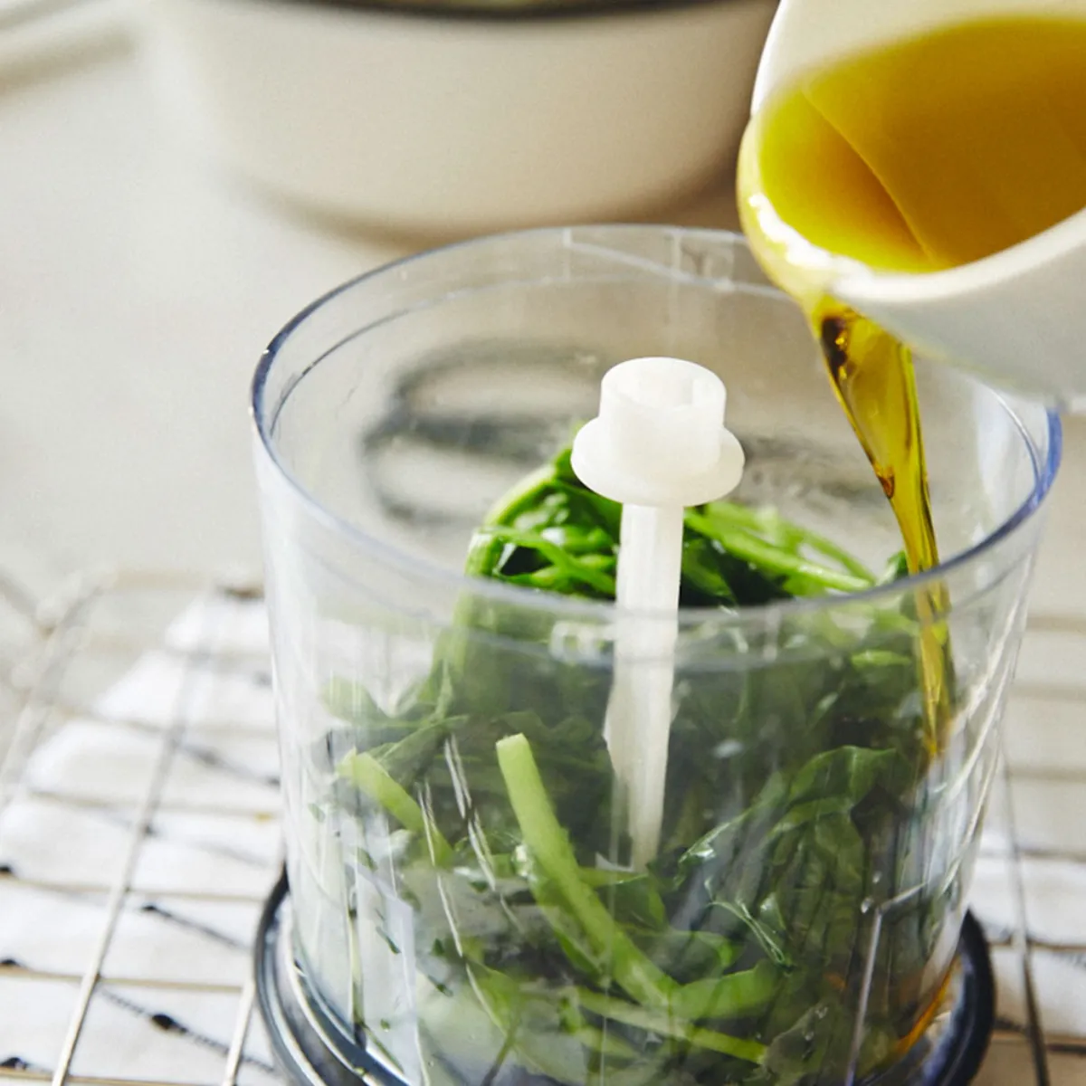 Versare l'olio in un robot da cucina contenente erbe aromatiche fresche, preparando gli ingredienti da frullare sul piano di lavoro della cucina.