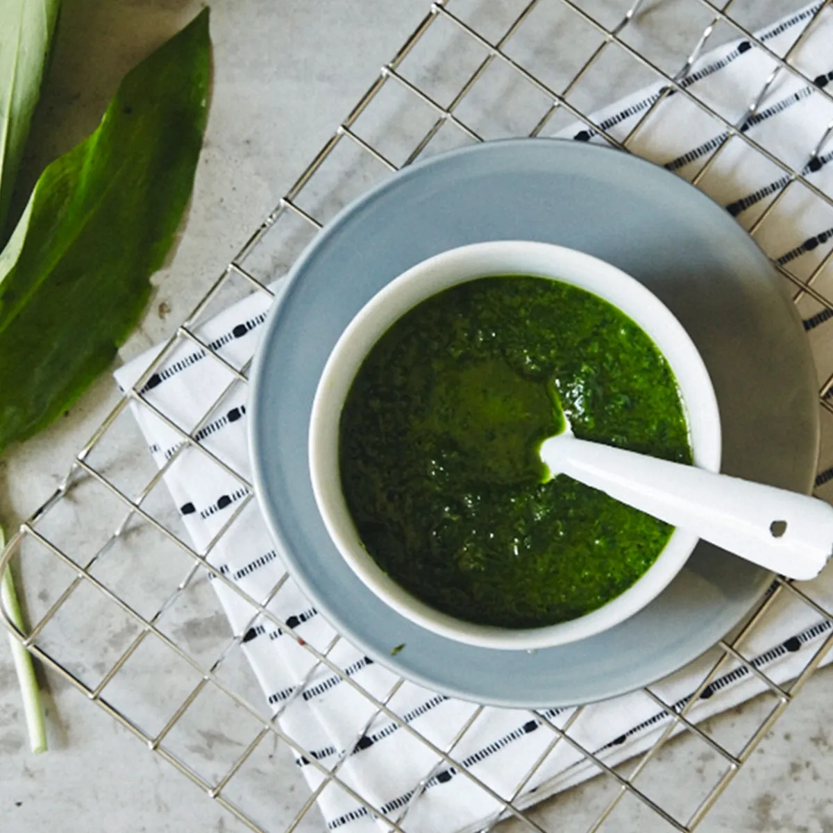 Ciotola di salsa alle erbe con un cucchiaio bianco, appoggiata su un piatto grigio e una tovaglia a quadretti, accanto a foglie verdi fresche su una superficie grigia.