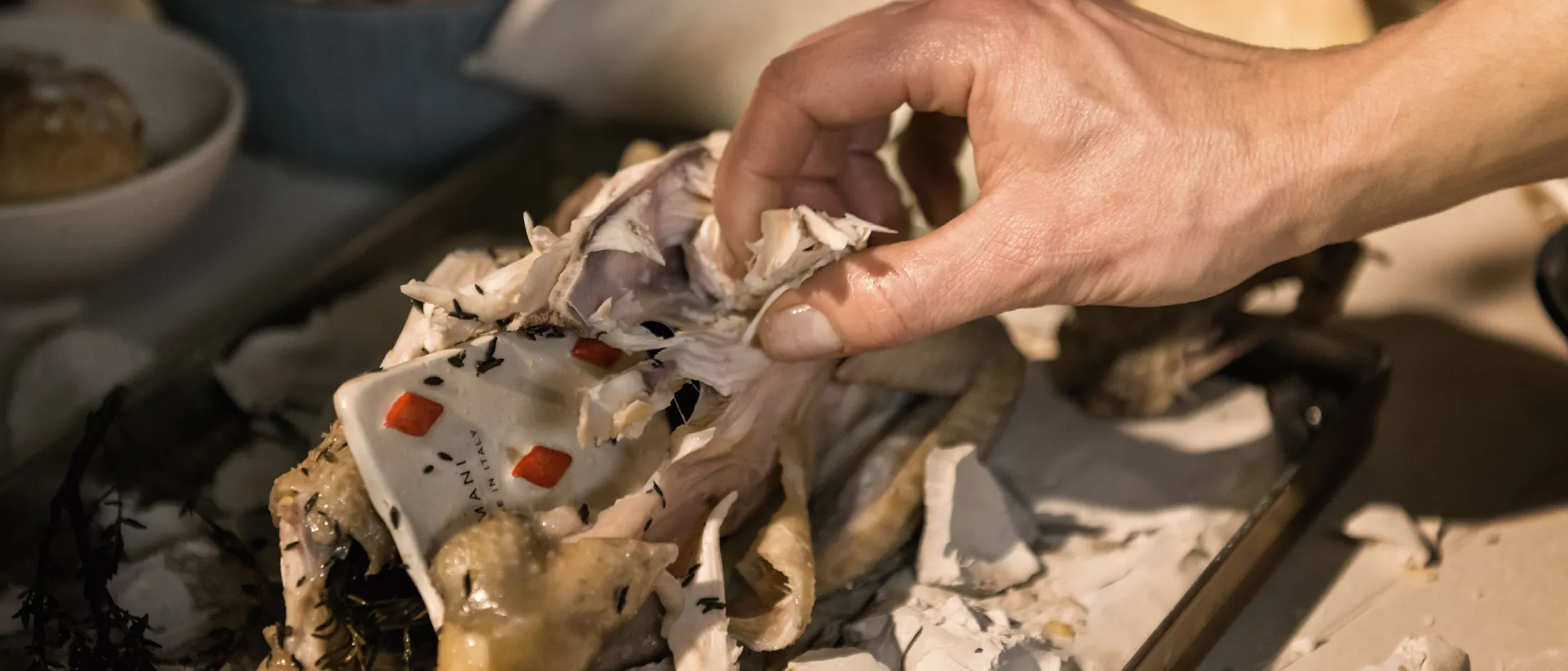 Una mano che solleva una carta da gioco traforata avvolta in un pollo arrosto su un vassoio, con erbe aromatiche e pane sullo sfondo.