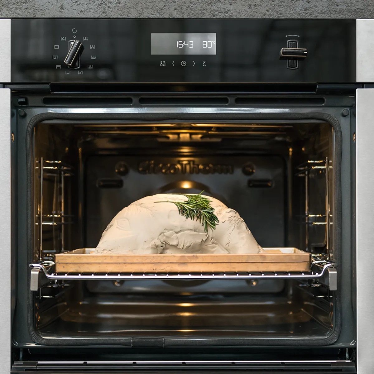 Un moderno forno in acciaio inossidabile con all'interno un arrosto coperto, guarnito con rosmarino fresco, impostato a 80 °C, mentre l'orologio digitale segna le 15:43.