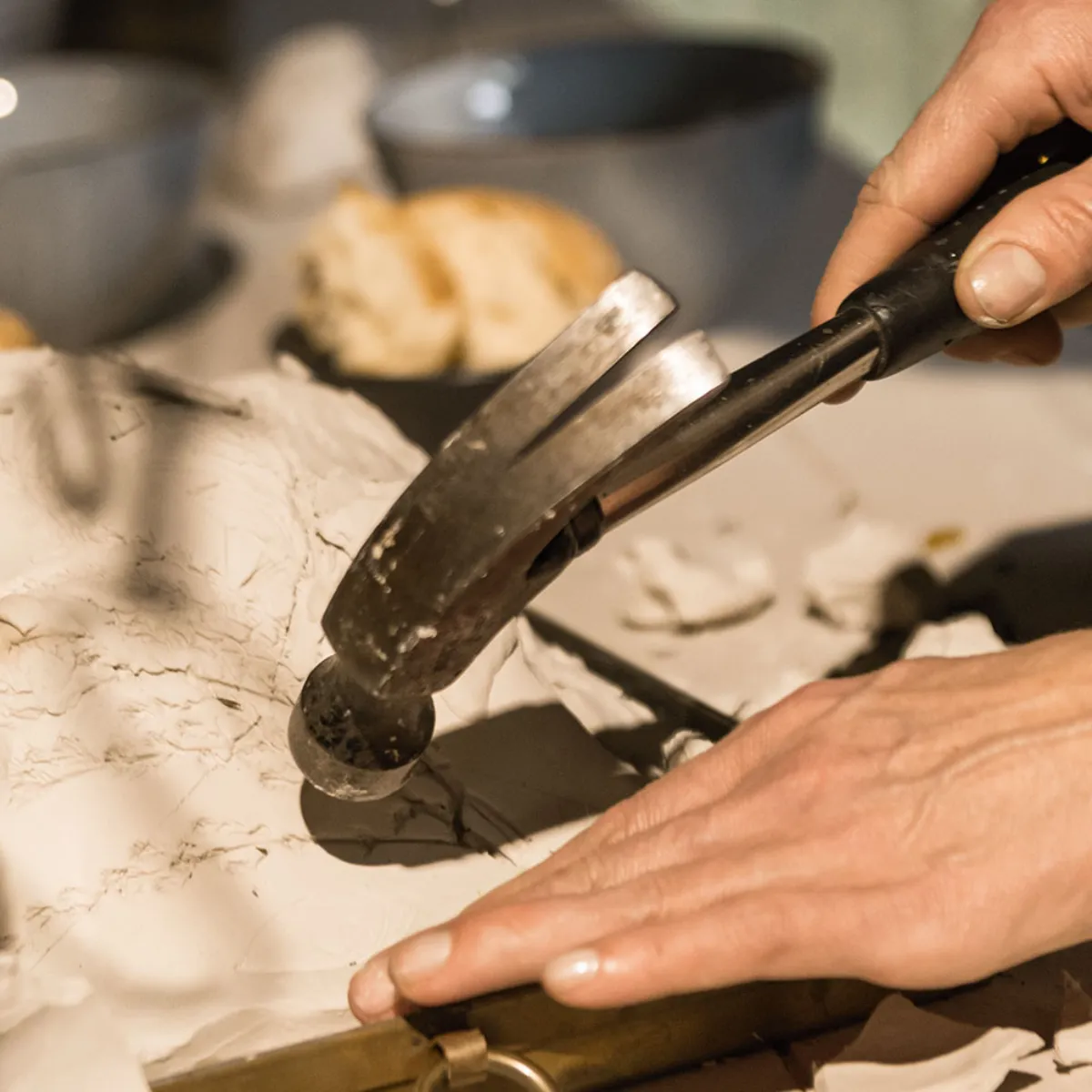 Mani che utilizzano un martello per scolpire o modellare un materiale bianco su un tavolo, con oggetti sfocati sullo sfondo, tra cui ciotole e pane.