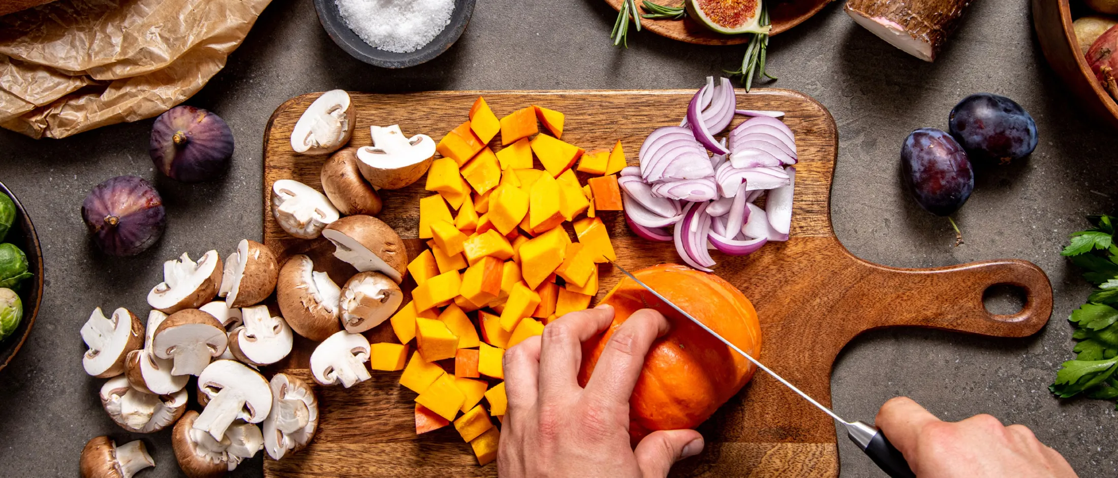 Mani che tagliano una zucca a cubetti su un tagliere di legno con funghi affettati cipolla rossa a fette e altri ingredienti intorno