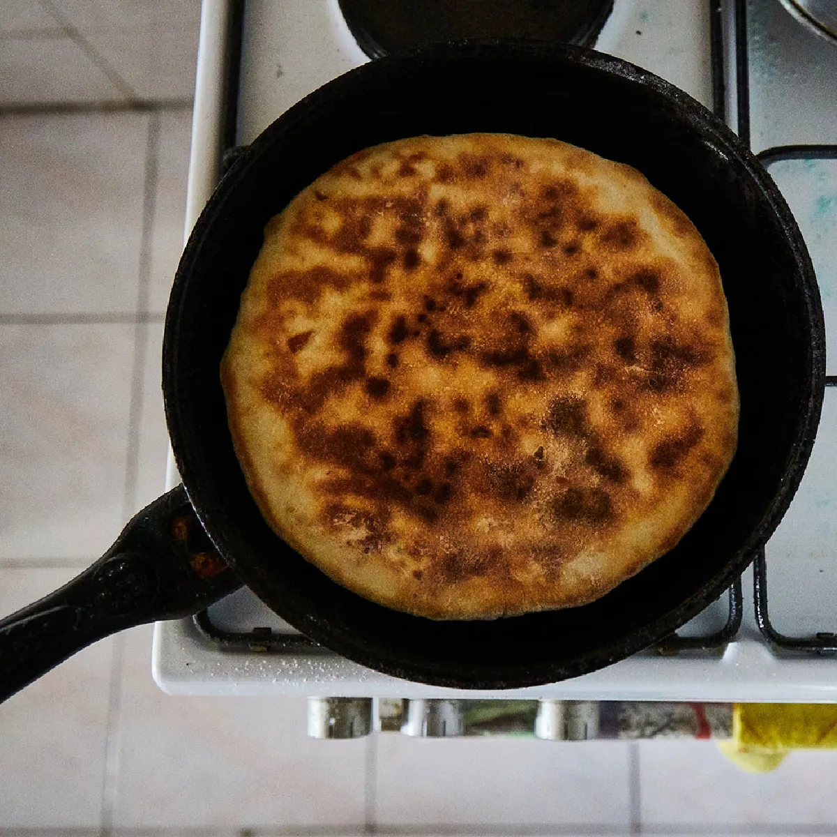 Vista dall'alto di un pane piatto dorato e ben cotto in una padella nera sul fornello a gas.