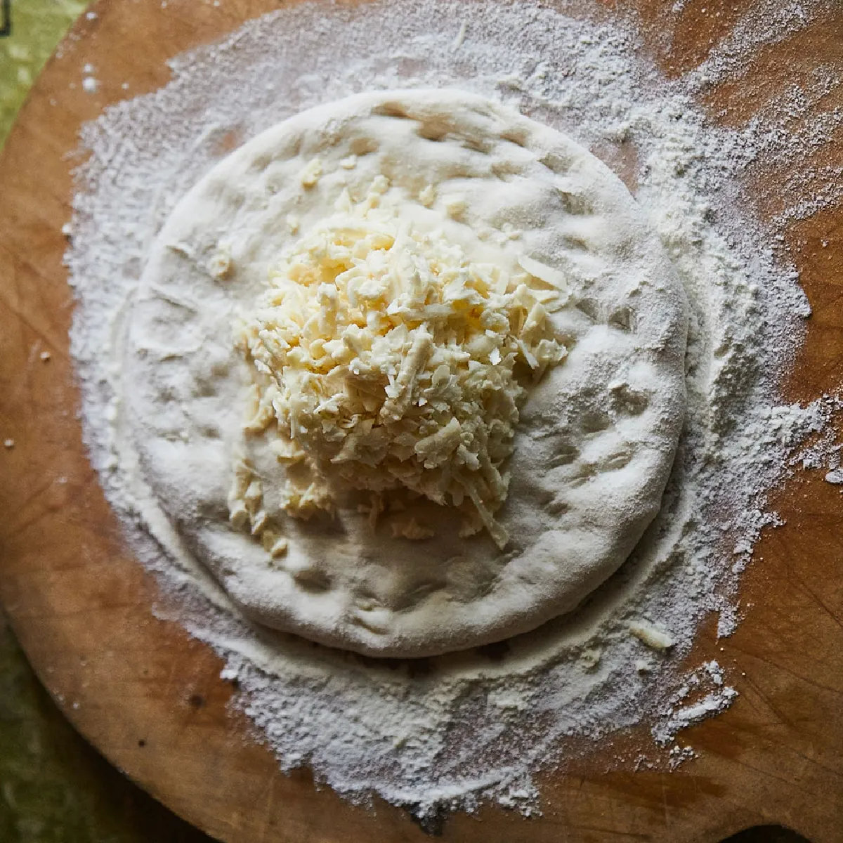 Impasto rotondo al centro di un tagliere di legno infarinato con una montagnetta di formaggio grattugiato sopra; sullo sfondo una tovaglia verde e una mano parzialmente visibile a sinistra.