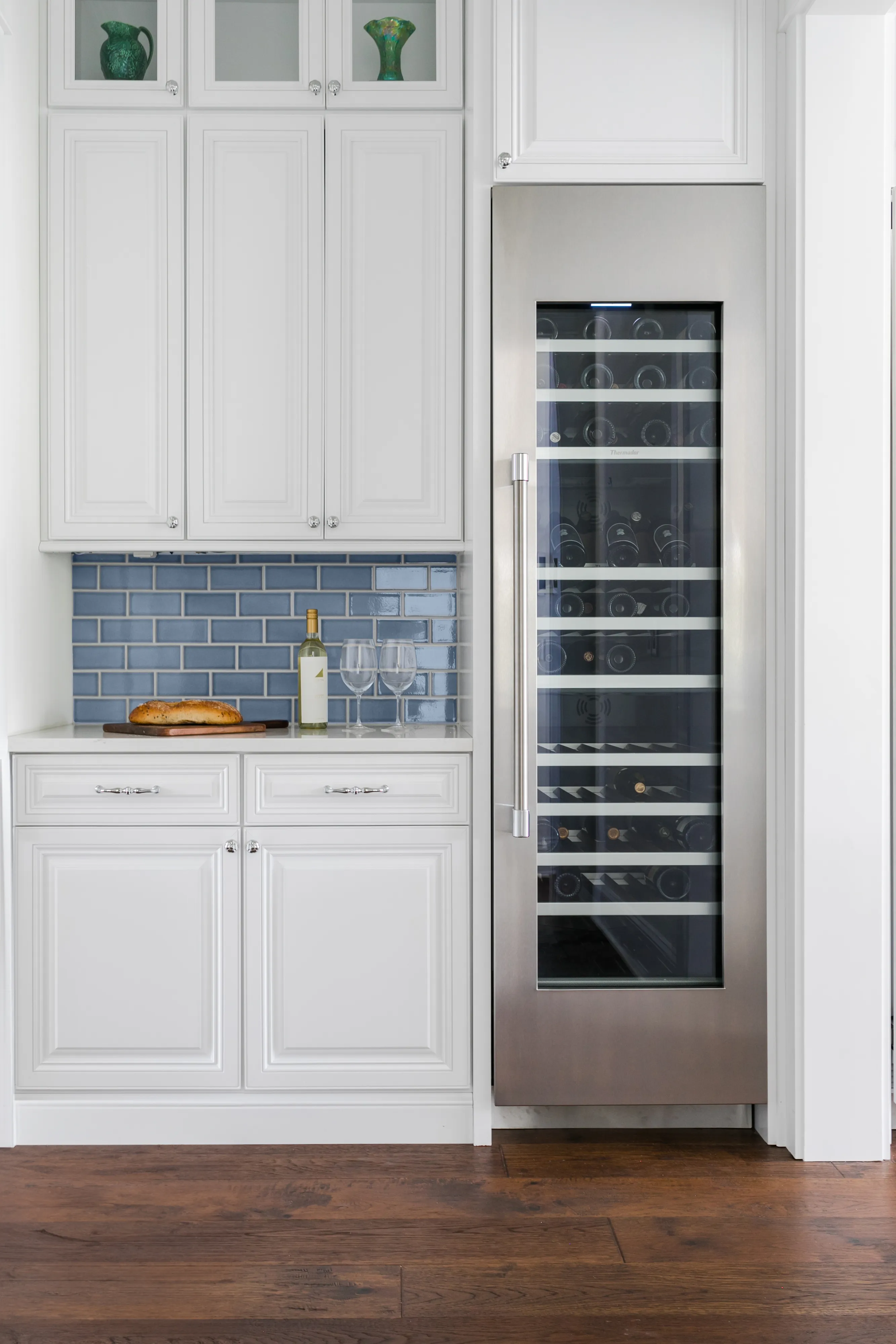 White kitchen cabinets with blue tile backsplash, a bottle of white wine, two wine glasses, a loaf of bread, and a stainless steel wine fridge.