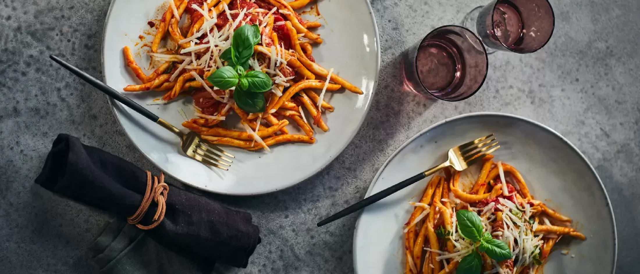Piatti di pasta con salsa di pomodoro, guarniti con foglie di basilico fresco, accompagnati da bicchieri di vino.