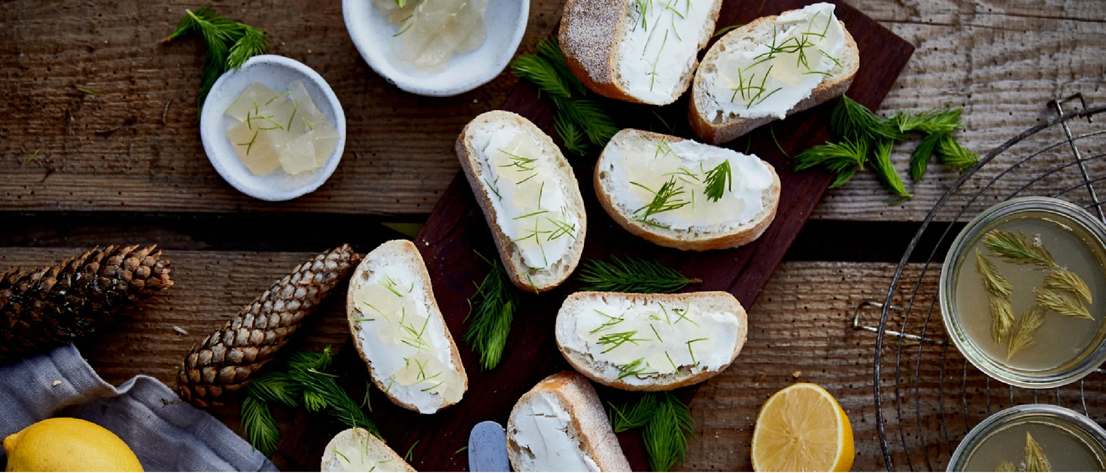 Fette di pane con formaggio e punte di abete su un tagliere scuro, con ciotole, pigne e mezzo limone.