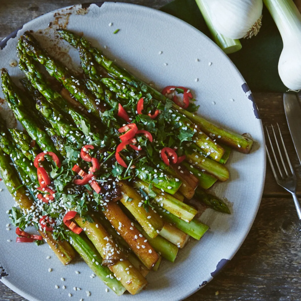 Asparagi saltati con semi di sesamo e fettine di peperoncino rosso su piatto bianco con posate e cipolla su tavolo di legno.