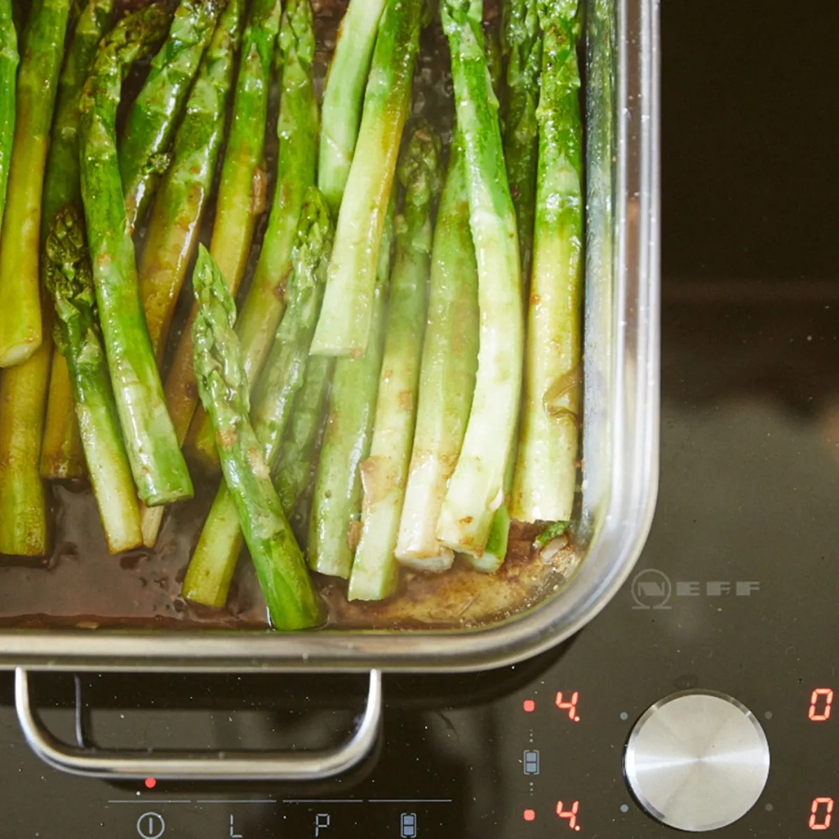 Asparagi verdi disposti in una teglia rettangolare trasparente su piano cottura nero con manopola argentata e display digitale rosso.