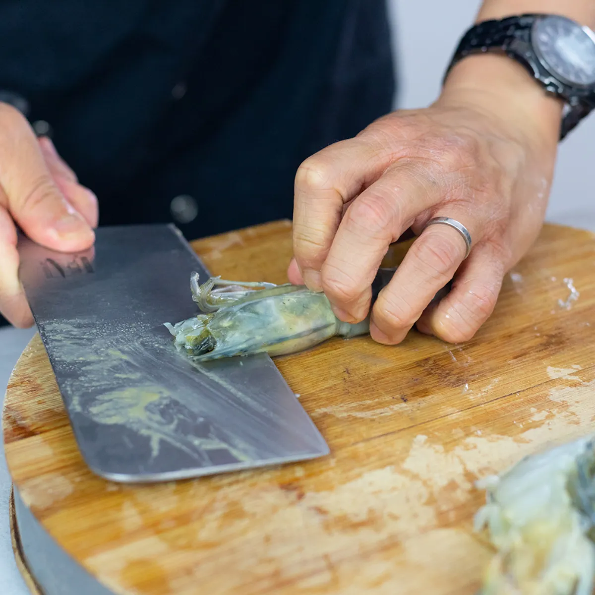 Mani che tagliano gamberi crudi su un tagliere rotondo in legno con un coltello largo.
