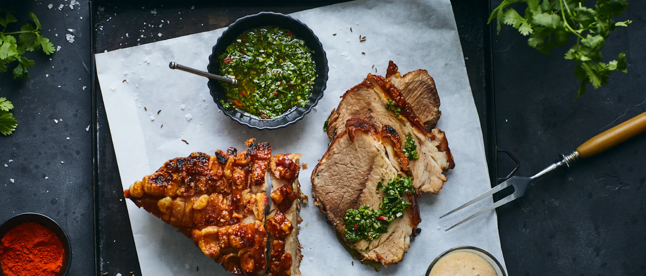 Carne alla griglia, salsa di chimichurri, verdure arrosto, spezie e utensili da cucina su uno sfondo scuro.