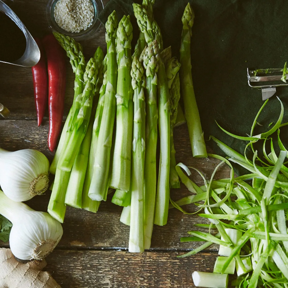 Asparagi verdi interi e pelati, cipollotti, peperoncini rossi, coriandolo, zenzero, spezie e salse disposti su un piano di legno con un coltello e un pelapatate.