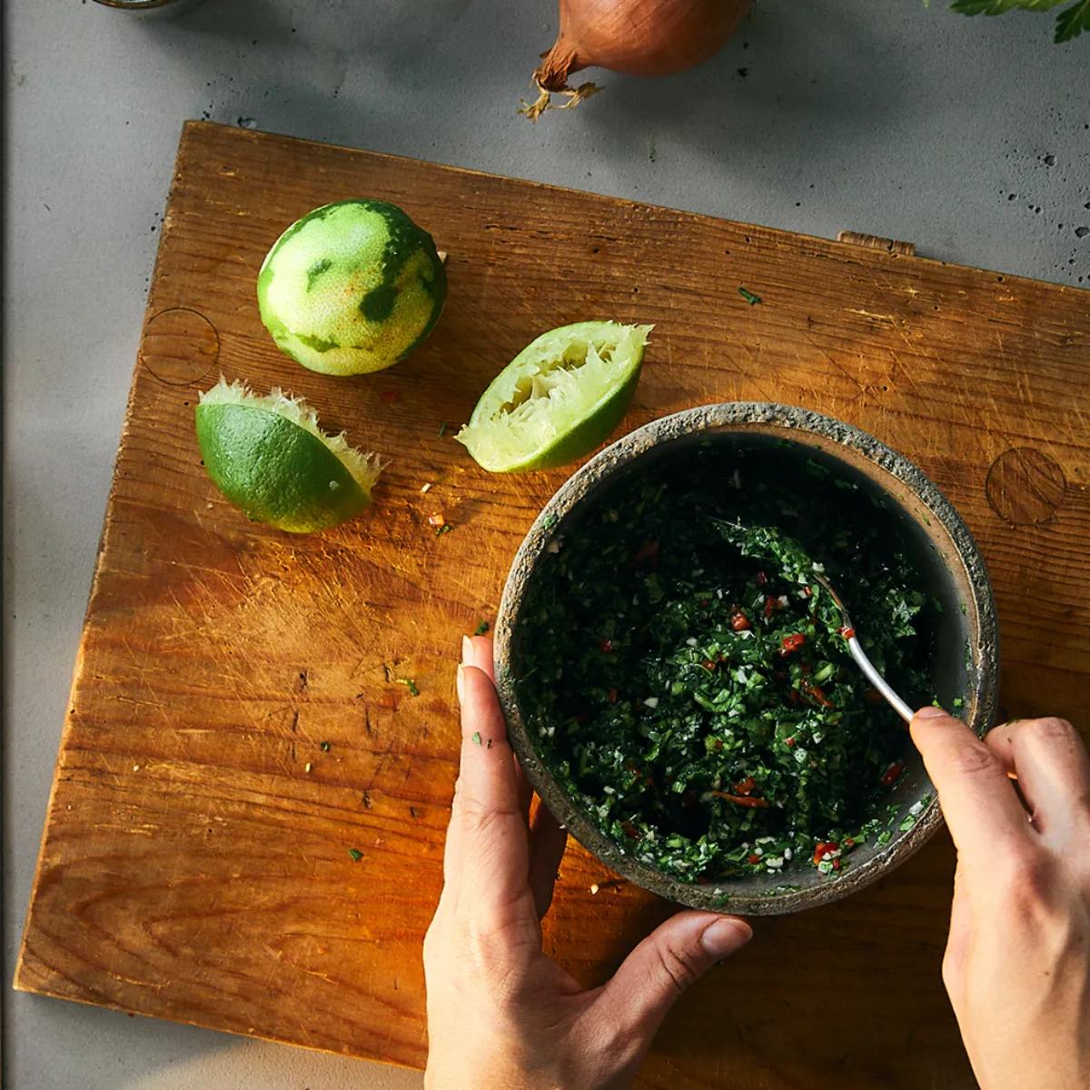 Mani che mescolano una salsa verde in una ciotola di pietra su un tagliere di legno con lime tagliati accanto.