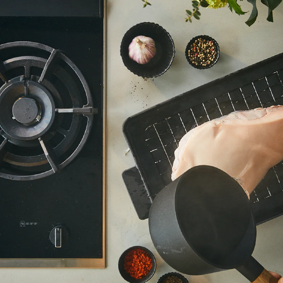 Pezzo di carne cruda su griglia con pentola nera che versa liquido sopra, accanto fornello a gas e ciotole con spezie e aglio.