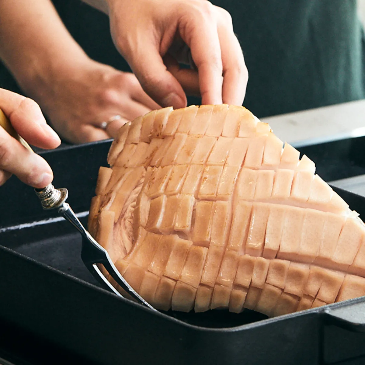 Mano che tiene una forchetta e un coltello mentre incidono la superficie di un pezzo di carne con tagli a griglia in una teglia nera.