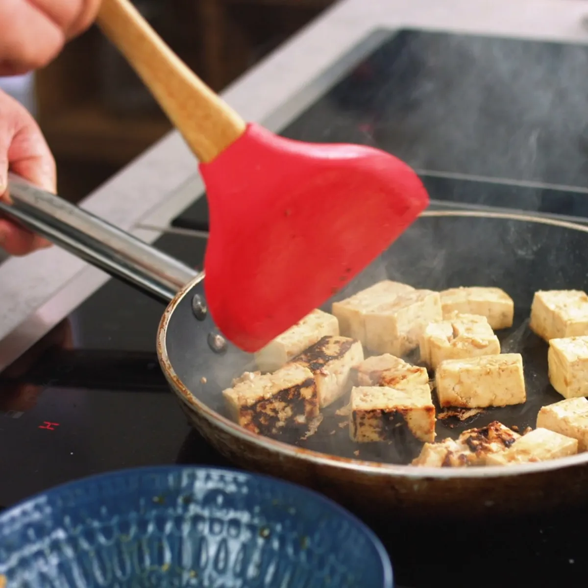 Cubetti di tofu che cuociono in padella con spatola rossa e manico in legno in mano.