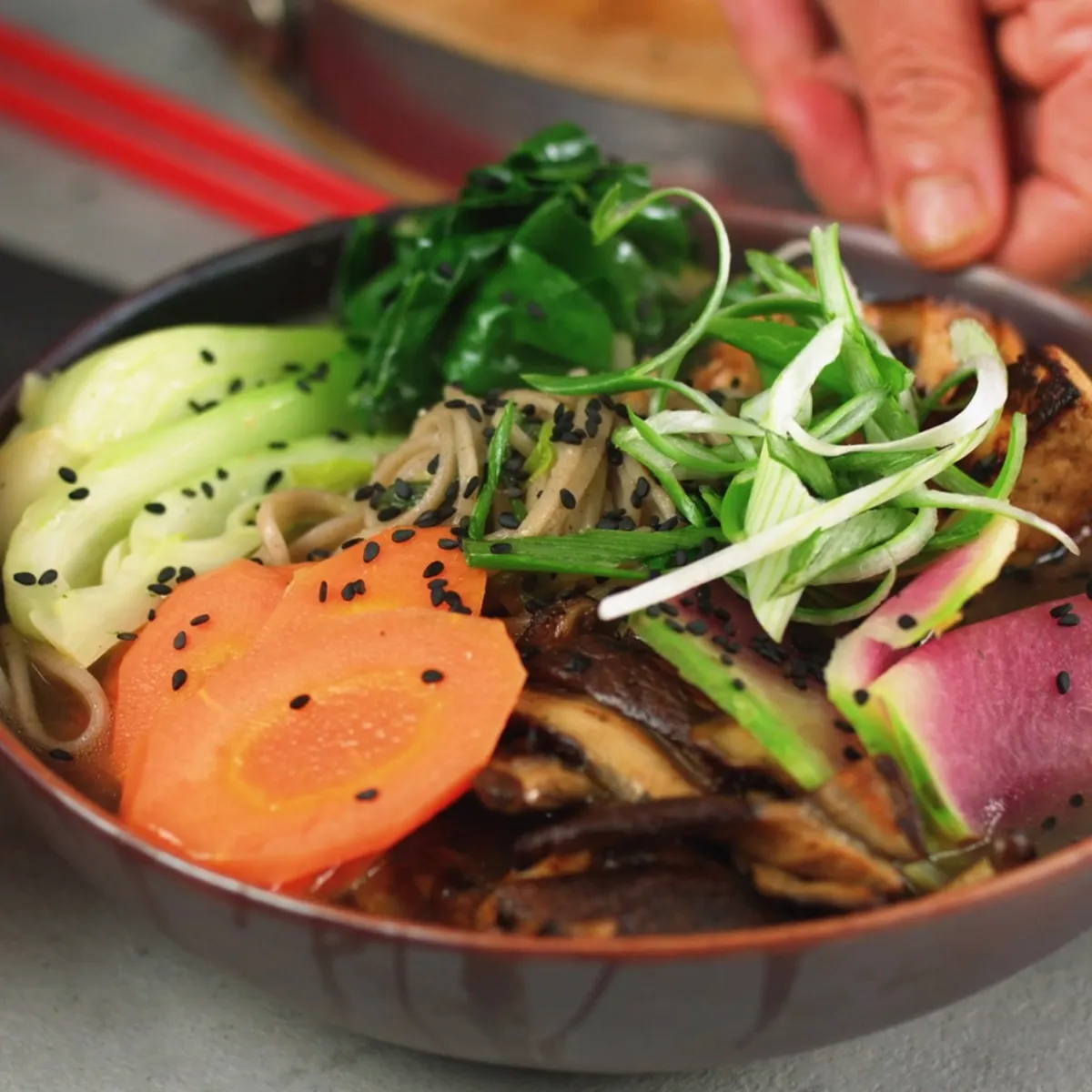 Ciotola con noodles soba, fette di carota, cetriolo, ravanello, funghi, spinaci e germogli, mani che la tengono.