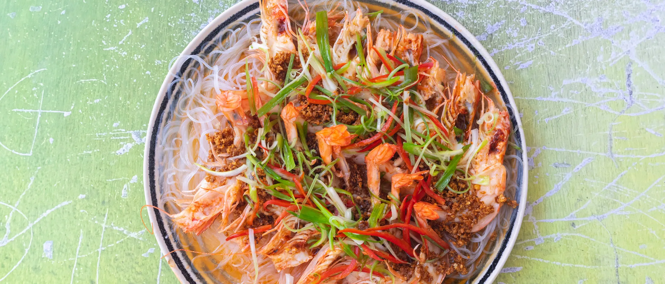 Piatto visto dall'alto con vermicelli di riso, gamberi, fette di peperoncino rosso e strisce di cipollotto verde su sfondo verde chiaro.