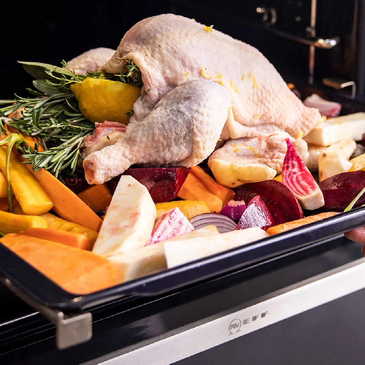 Un pollo intero crudo ripieno di erbe aromatiche e limone, adagiato su un vassoio con verdure colorate, viene messo in forno per la cottura