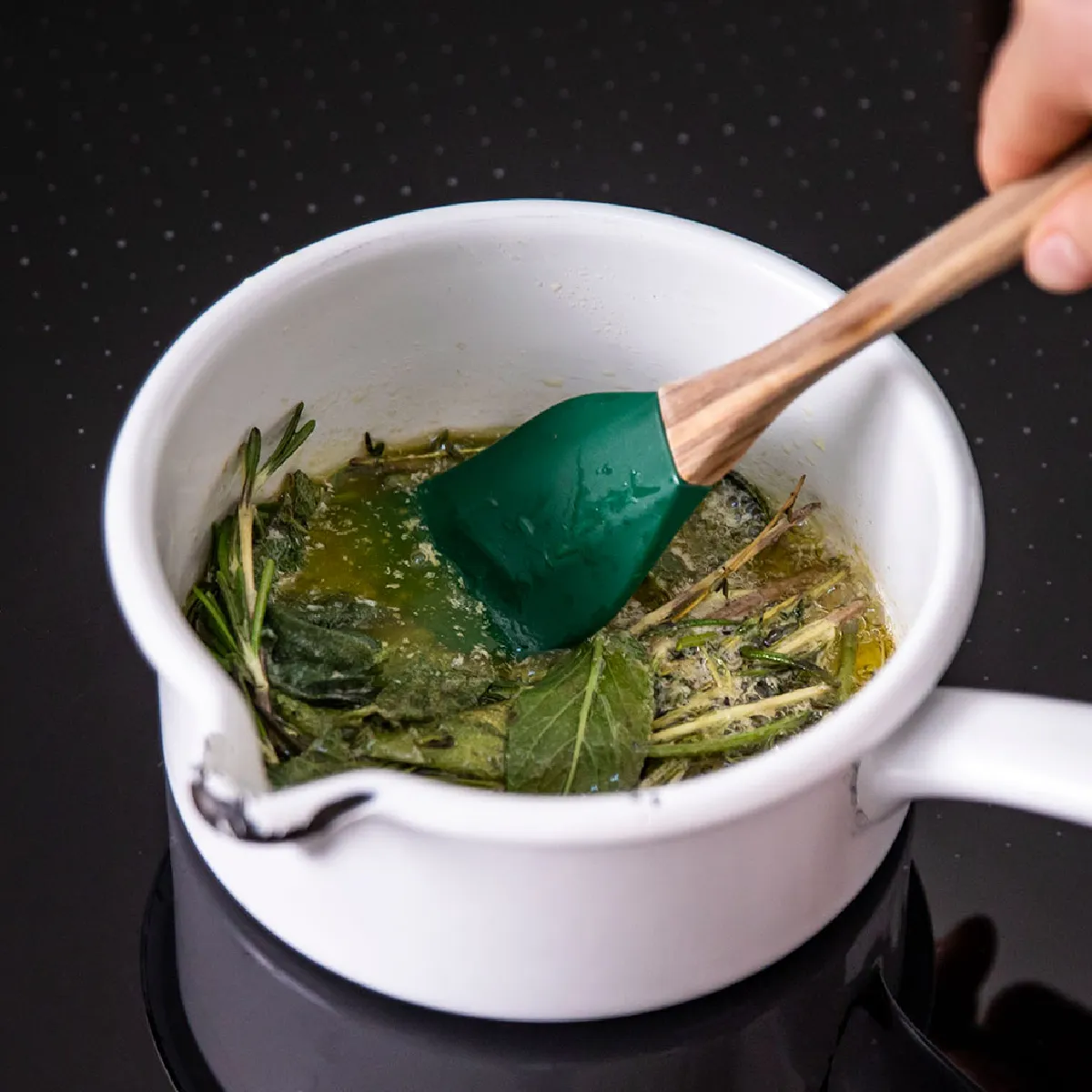 Mano che mescola erbe aromatiche in olio dentro una casseruola bianca su piano cottura nero con spatola verde dal manico in legno