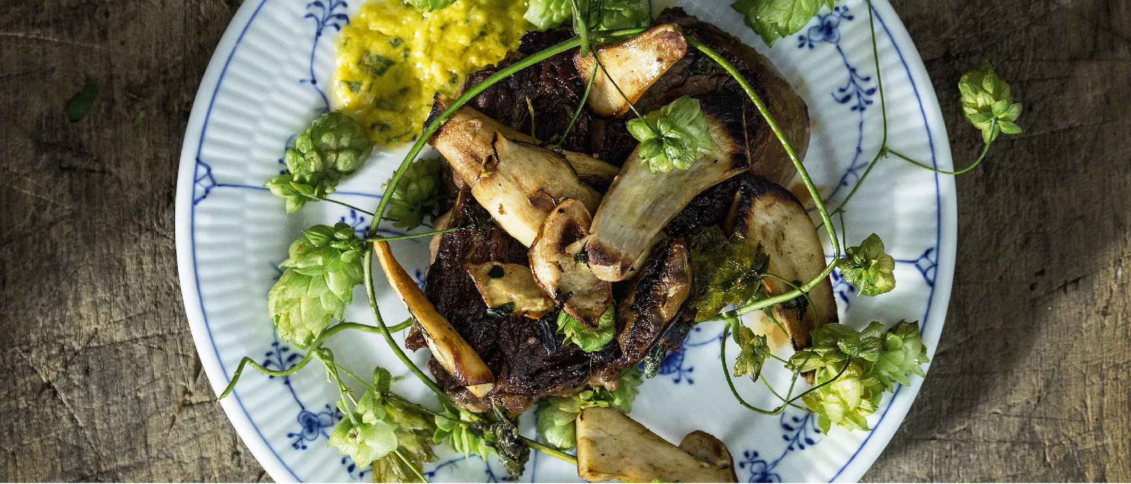 Piatto con funghi grigliati, fiori di luppolo verdi e una salsa gialla su un vassoio rustico di legno