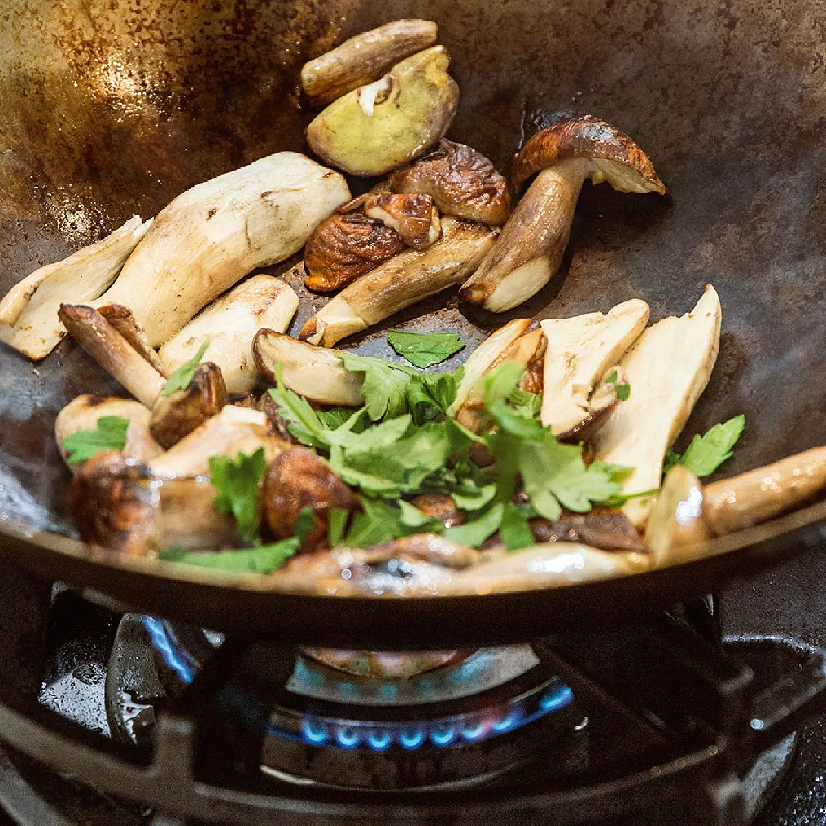Funghi ed erbe aromatiche fresche saltati in un wok rustico di metallo su un fornello a gas.