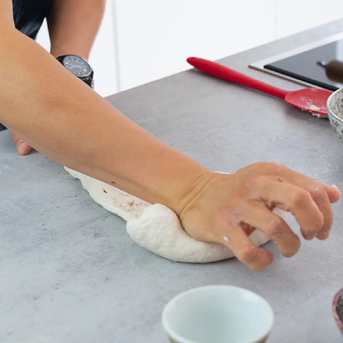 Mani che impastano la pasta su un bancone della cucina, con ciotole e utensili nelle vicinanze, durante la preparazione del pane