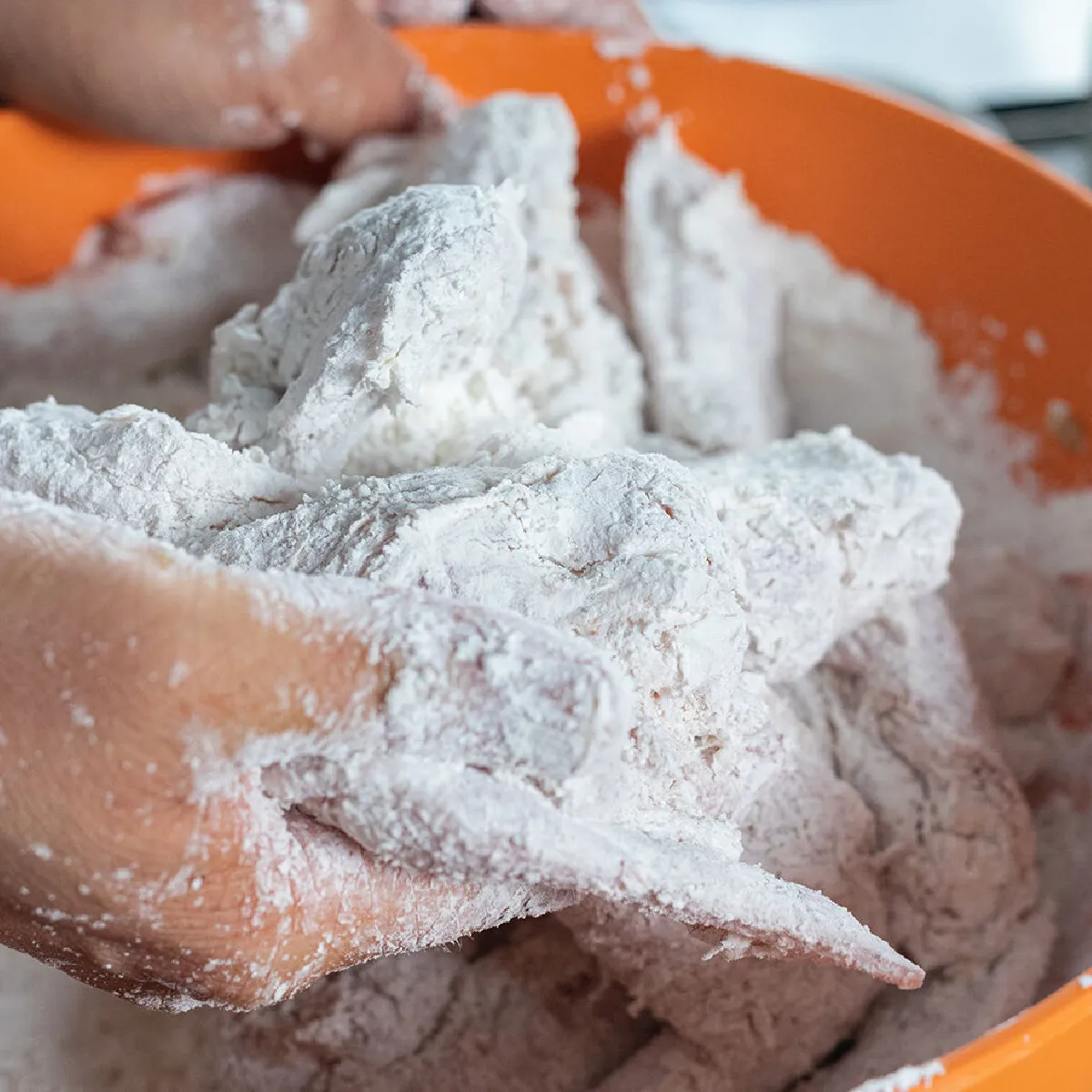 Mani che ricoprono pezzi di pollo con farina in una grande ciotola arancione; sullo sfondo una tazza e utensili da cucina sfocati.