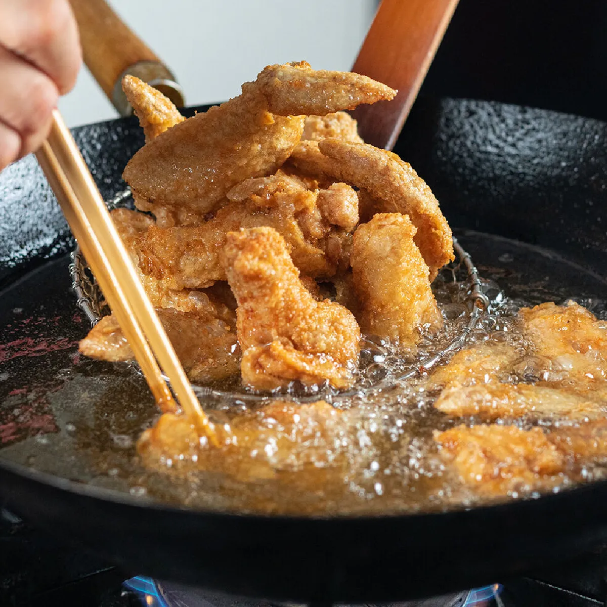 Pezzi di pollo impanati che frigtono in olio bollente in un wok nero su un fornello, con una mano che usa bacchette e una spatola per sollevare i pezzi mentre l'olio sfrigola.