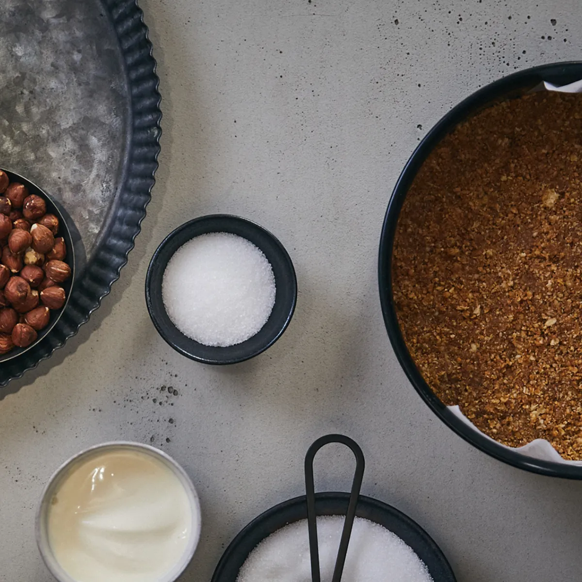 Base di biscotti pressata in stampo rotondo con piccoli contenitori di nocciole, zucchero e crema intorno