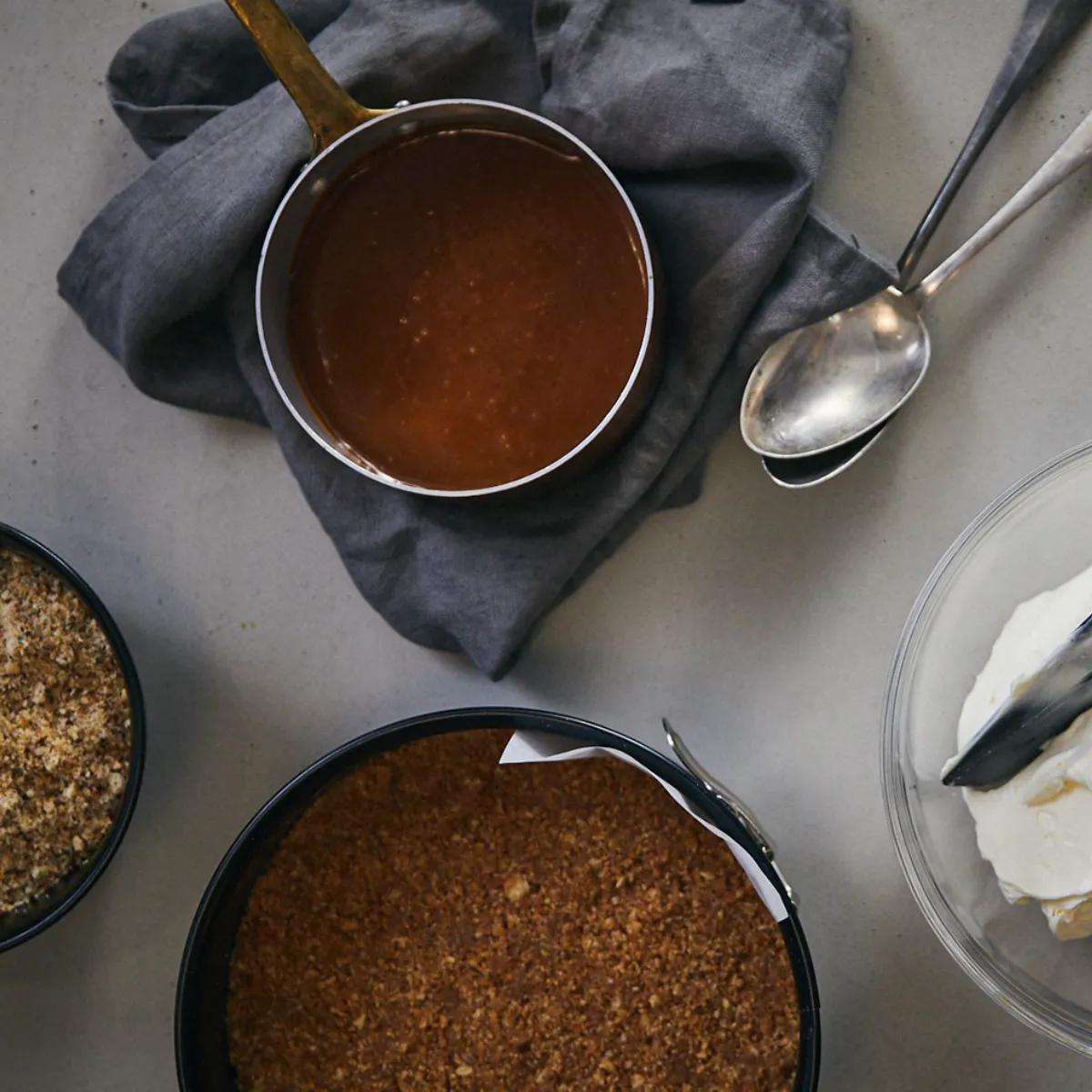 Quattro contenitori con ingredienti per torta: miscela di biscotti tritati, salsa marrone in pentolino, base di torta in stampo apribile e crema bianca in ciotola con spatola