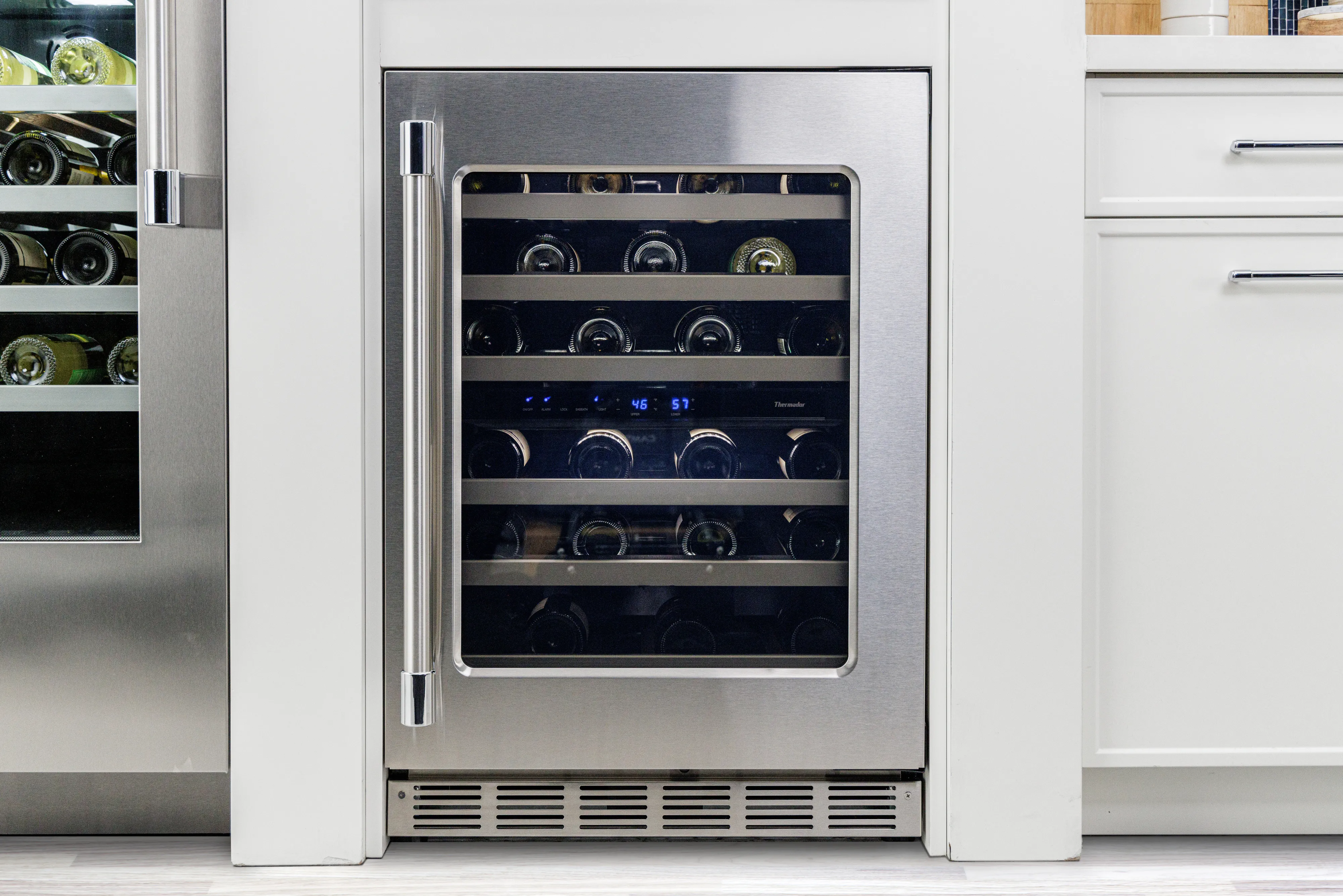 Built-in stainless steel wine cooler with glass door, holding multiple wine bottles inside a modern white kitchen.