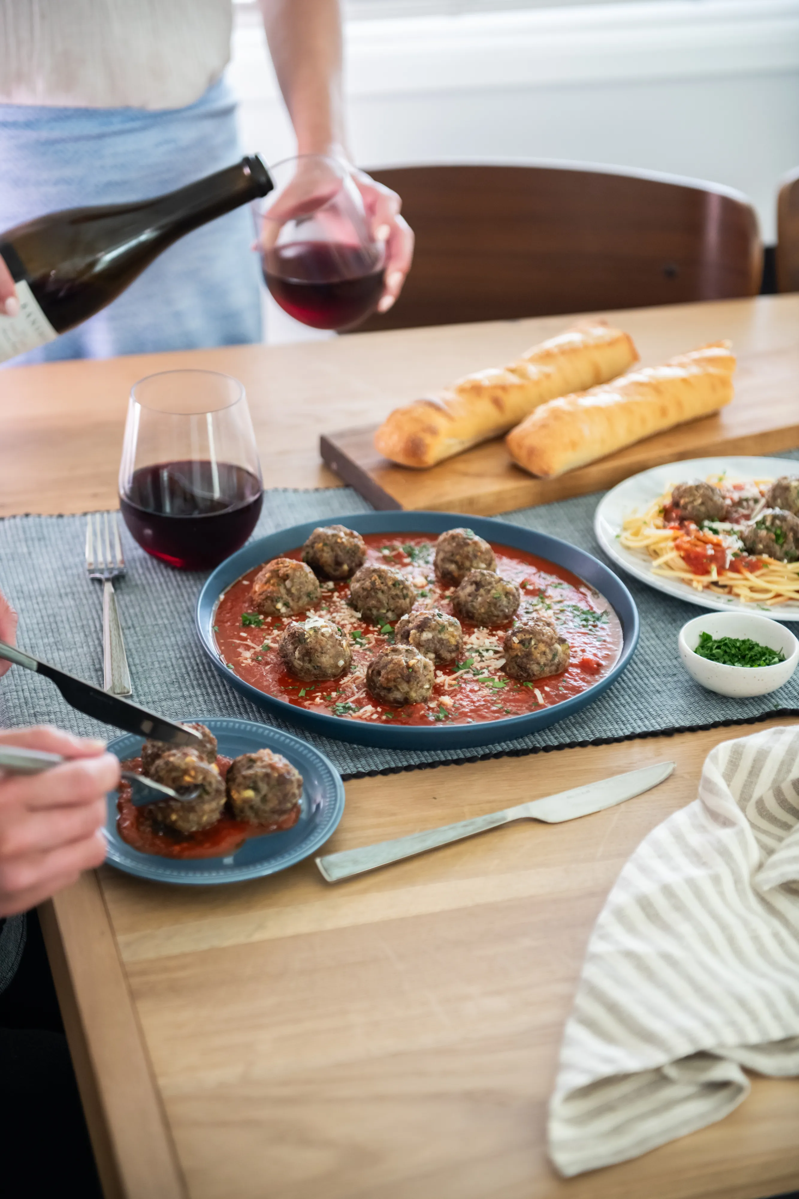 Person serving meatballs with spaghetti and bread on a wooden table, with glasses of red wine being poured.