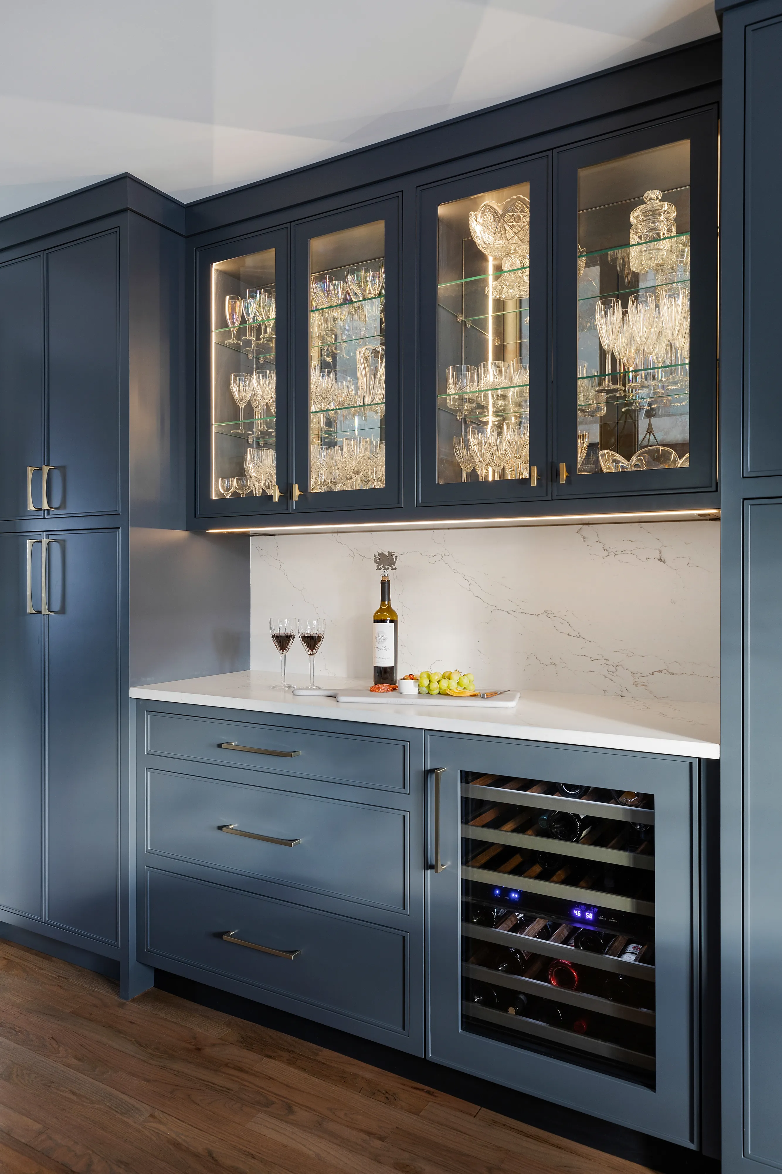 Modern blue kitchen cabinetry with glassware display, wine cooler, marble backsplash, and countertop holding wine glasses, bottle, and grapes.
